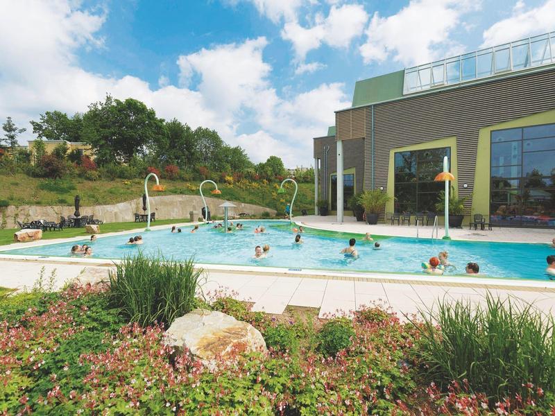 Piscina exterior con personas, jardín y edificio de hotel bajo cielo soleado.