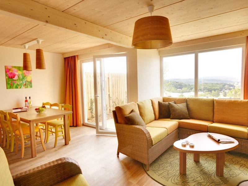 Sala de estar luminosa con sofá beige, mesa redonda y comedor junto a ventana con vista al campo.