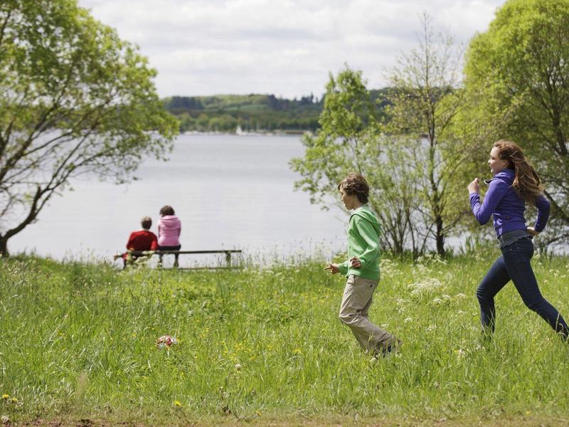 Niños juegan en un prado junto al lago mientras dos personas están sentadas en un banco.