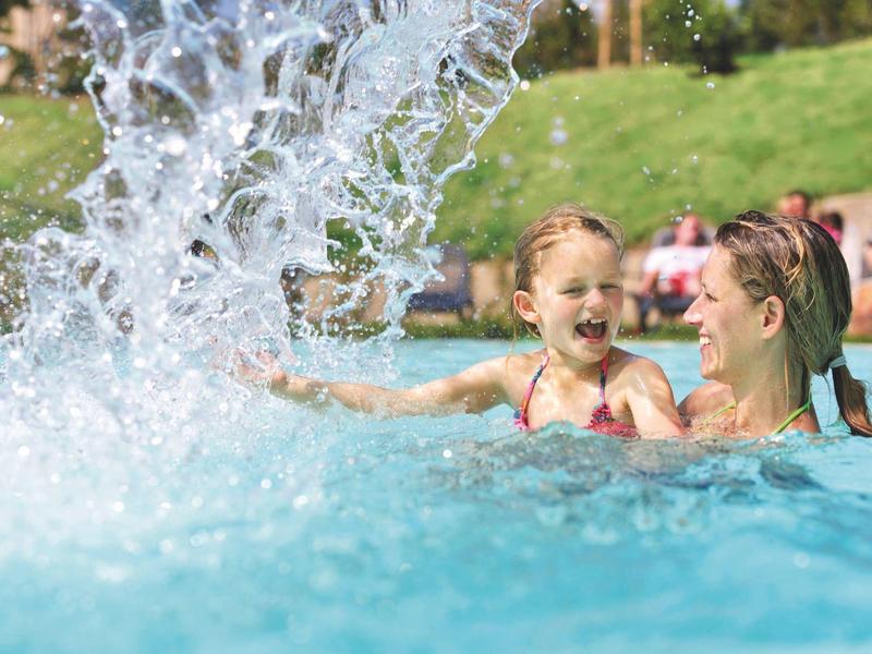 Madre e hijo juegan riendo en la piscina con un chorro de agua de fondo.
