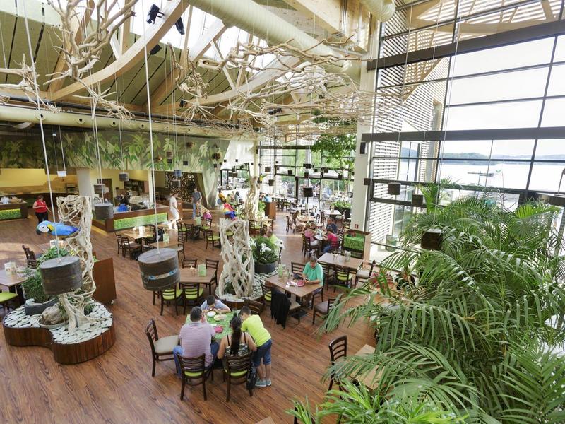 Amplio restaurante interior con techo de vidrio, plantas decorativas y clientes sentados en mesas redondas.