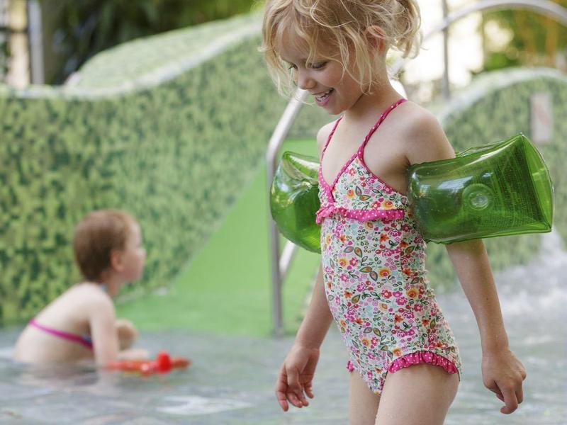 Dos niños juegan en una piscina poco profunda con agua y un tobogán verde al fondo.