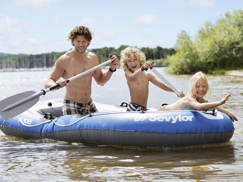 Tres niños reman en un bote inflable azul en agua tranquila con paisaje verde en la orilla.