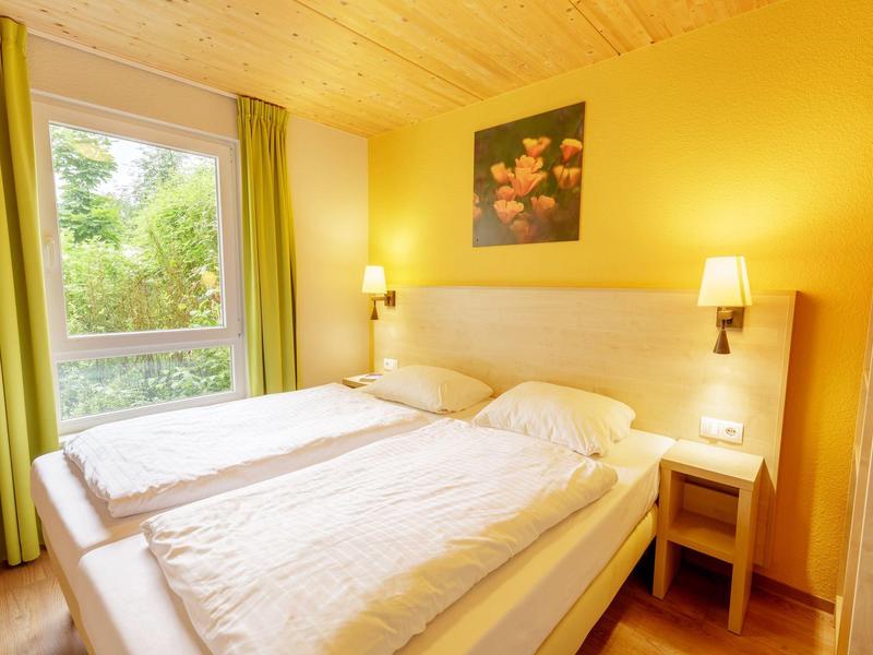 Dormitorio luminoso con dos camas individuales, techo de madera y ventana grande con vista al verde.