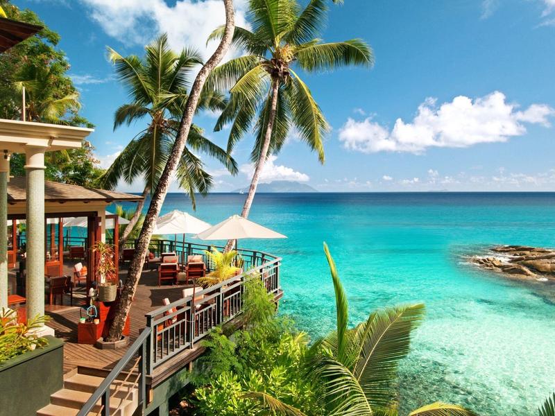 Bar sulla spiaggia tropicale con terrazza e palme accanto ad acqua turchese chiara.
