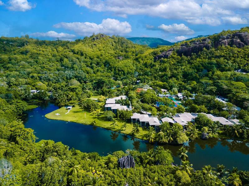 Complexe hôtelier au bord d'une rivière entourée de collines vertes et de montagnes boisées sous un ciel bleu.