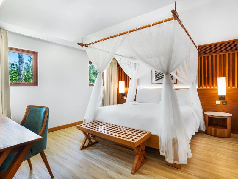 Chambre d'hôtel lumineuse avec lit à baldaquin, meubles en bois et fenêtre avec rideaux.