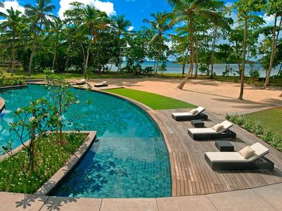 Pool mit Liegen, Palmen und Blick auf ruhige Lagune bei klarem Himmel.