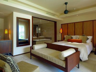 Chambre d'hôtel moderne avec grand lit, banc, lampes et espace salon avec coussins.