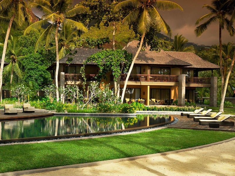 Complex tropical avec piscine incurvée, transats, palmiers et végétation luxuriante au crépuscule.