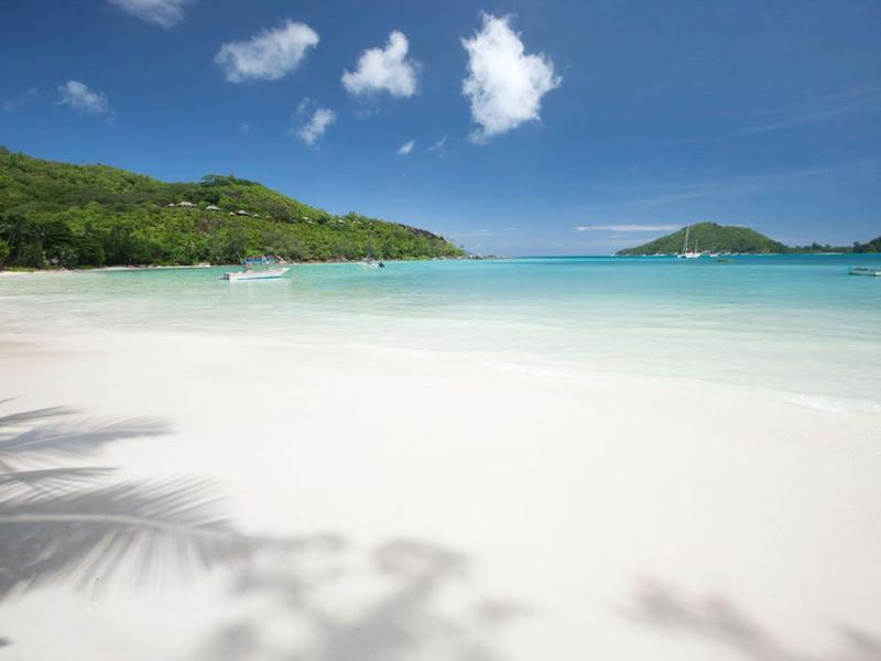 Plage de sable blanc immaculé avec eau turquoise claire et collines vertes sous un ciel bleu.