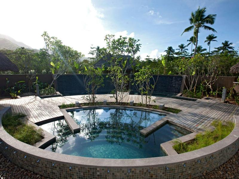 Piscine ronde à l'eau claire entourée d'arbres et d'une terrasse en bois sous un ciel bleu.