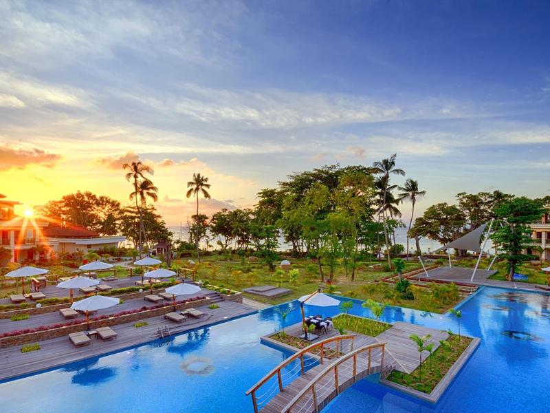 Ein großer Pool mit Holzliegen, Palmen und Sitzbereichen bei Sonnenuntergang im Resort.