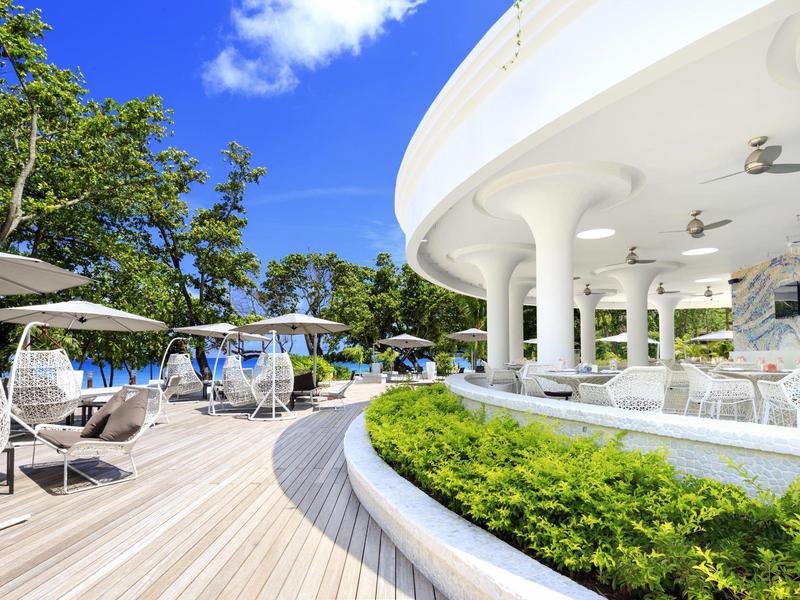 Moderner Außenbereich mit weißer Säulen-Terrasse, Holzboden, Pflanzen und Sitzgelegenheiten unter blauem Himmel.