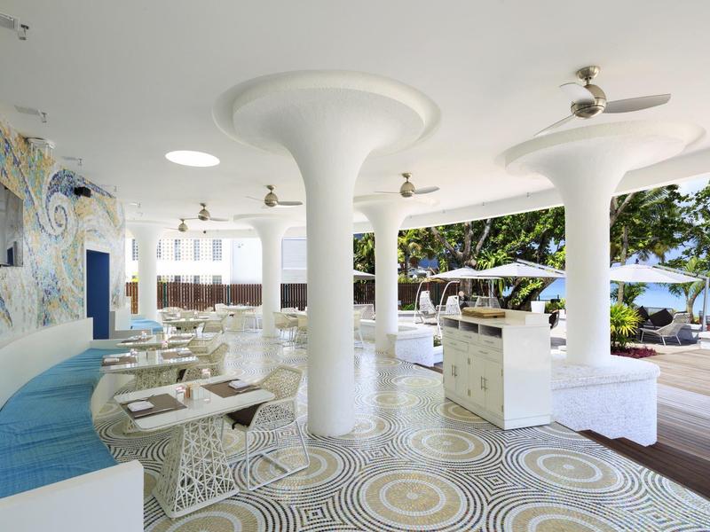 Moderner, offener Restaurantbereich mit weißen Säulen, gemustertem Boden und Blick auf Meer und Bäume.