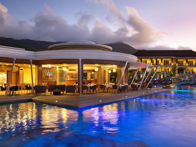 Modernes Hotel mit hellem Innenbereich, überdachten Terrassen und großem, blauen Pool bei Dämmerung.