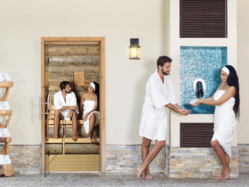 Paar in Bademänteln vor Sauna, Frau mit Handtuch, Wandlampen und Handtuchständer neben Tür.