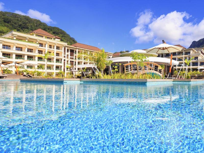 Großes Hotel mit mehreren Stockwerken, vor Bergkulisse, palmenumranktem Pool und Sonnenschirmen.