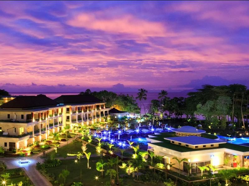 Hotelanlage bei Sonnenuntergang mit beleuchtetem Pool und üppiger Begrünung unter violettem Himmel.