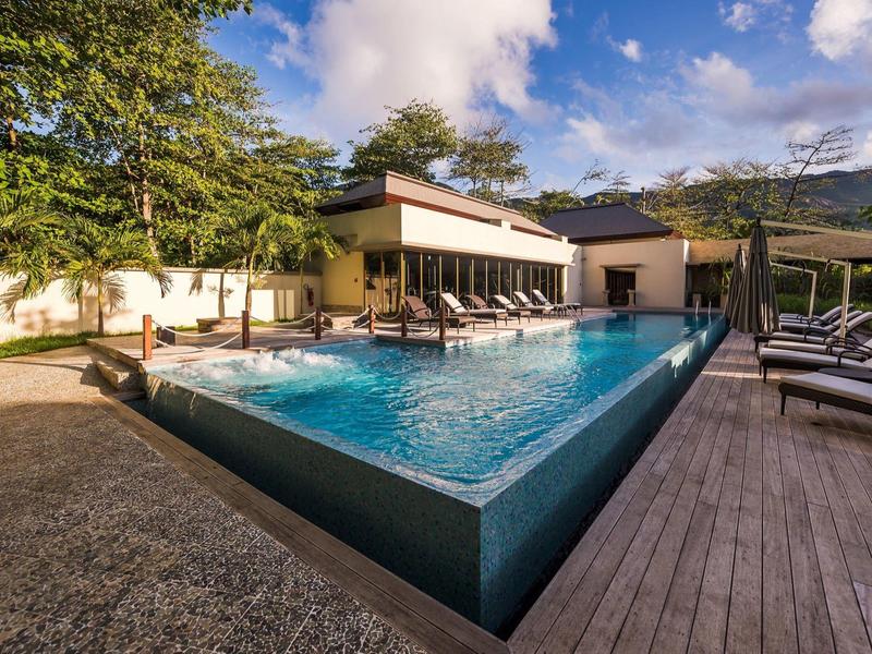Moderner Outdoor-Pool mit Liegestühlen, umgeben von Bäumen und Gebäuden bei blauem Himmel.