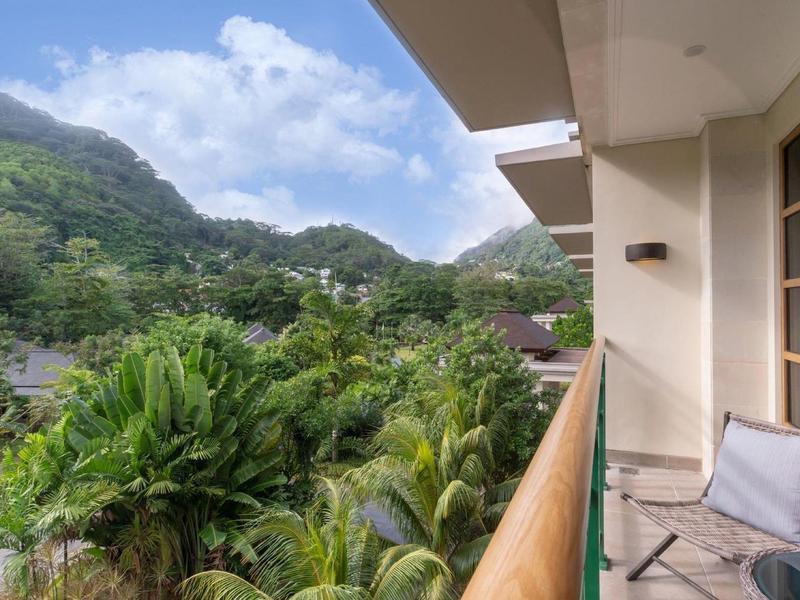 Balkon mit Stuhl, tropische grüne Pflanzen und bewachsene Hügel unter blauem Himmel.