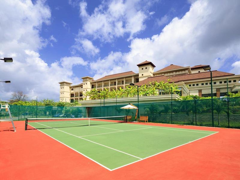 Tennisplatz mit grünem Spielfeld und rotem Rand vor mehrstöckigem Gebäude und blauem Himmel.
