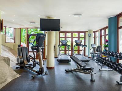 Hell beleuchteter Fitnessraum mit Trainingsgeräten und Hanteln neben großen Fenstern.