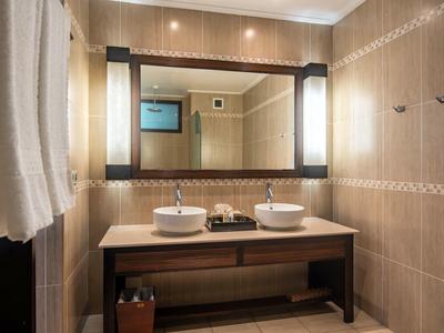 Área moderna de baño en hotel con doble lavabo, espejo grande iluminado y azulejos beige.