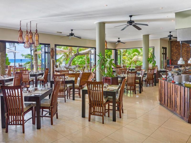 Restaurante espacioso con muebles de madera, ventiladores y grandes ventanas con vista a plantas tropicales.