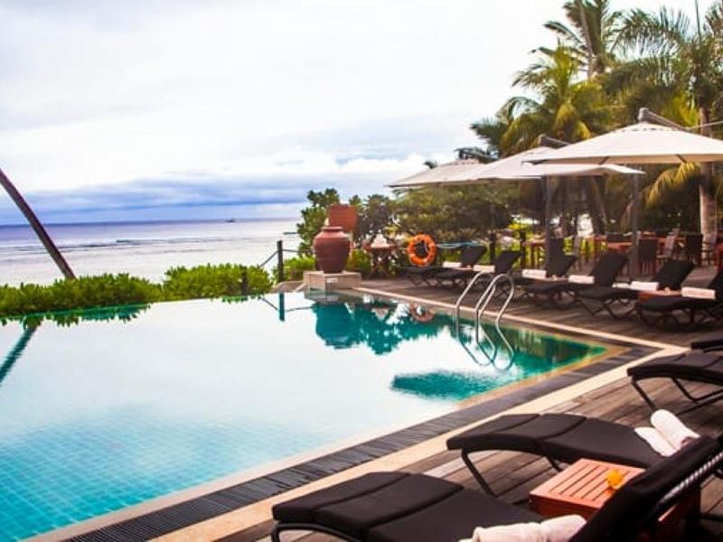 Piscina infinita con tumbonas y sombrillas junto a palmeras tropicales bajo cielo despejado.