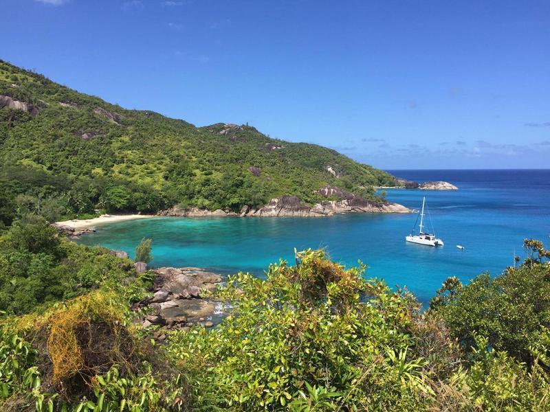 Bahía con agua turquesa, colinas verdes y un velero en el mar abierto.