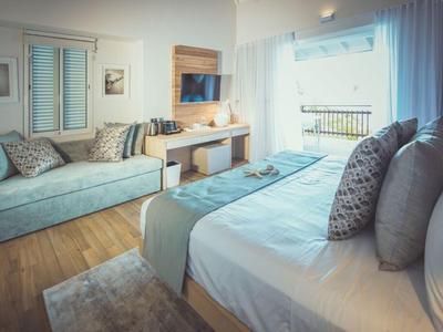 Světý hotelový pokoj s velkou postelí, pohovkou, dřevěnou podlahou a oknem se světlým závěsem.