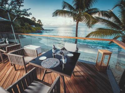 Balcon avec table à manger et chaises donnant sur une plage tropicale et l'océan avec des palmiers.