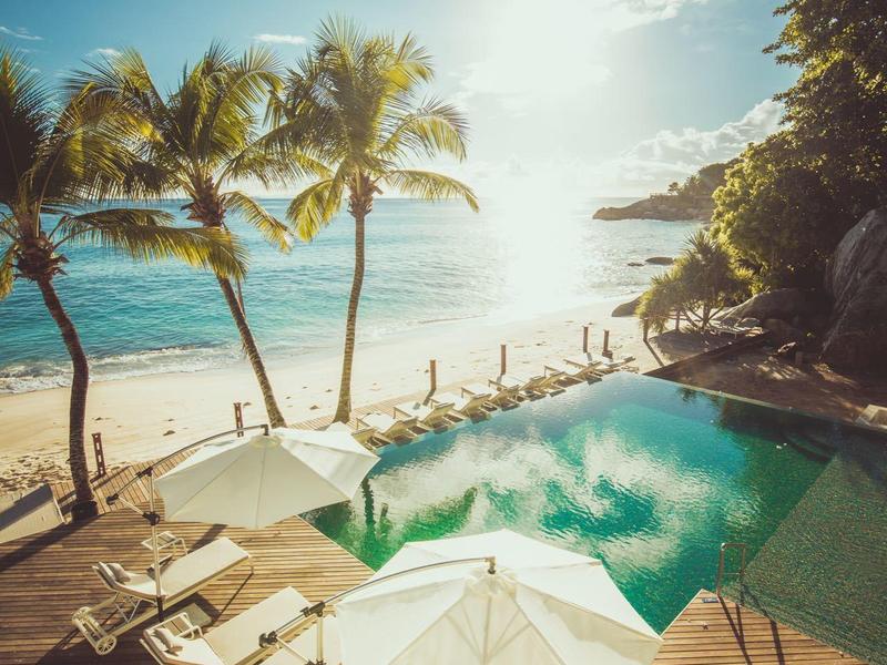 Bazén s slunečníky, lehátky a palmami na písečné pláži za slunečného počasí.