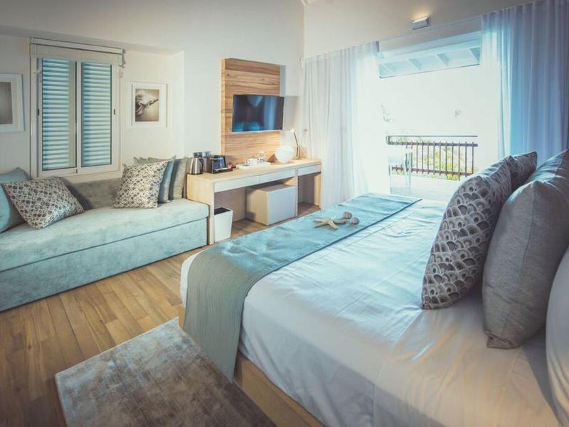 Světý hotelový pokoj s velkou postelí, pohovkou, dřevěnou podlahou a oknem se světlým závěsem.