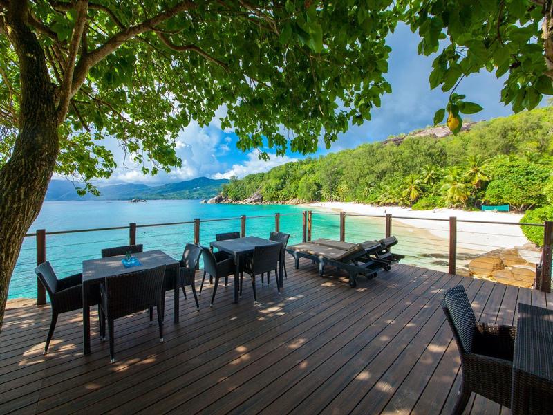 Terrazza con tavoli e sedie sull'acqua, circondata da alberi verdi e colline.