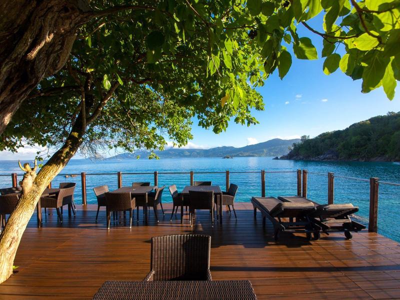 Terrazza con tavoli e sedie con vista sul mare sotto un albero in una giornata di sole.