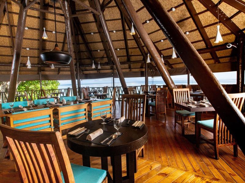 Área de restaurante abierta del hotel con muebles de madera y vista a la piscina y plantas tropicales.