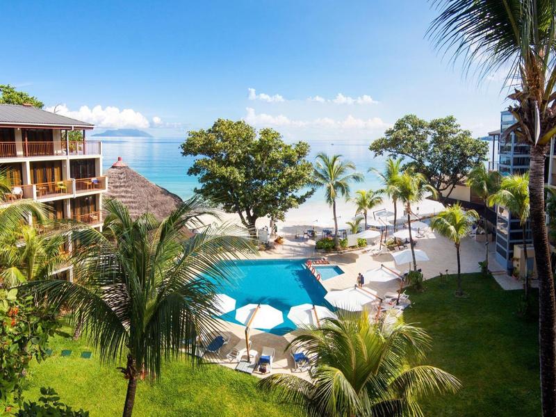 Complejo hotelero con piscina, sombrillas, palmeras y vista al mar bajo cielo despejado.