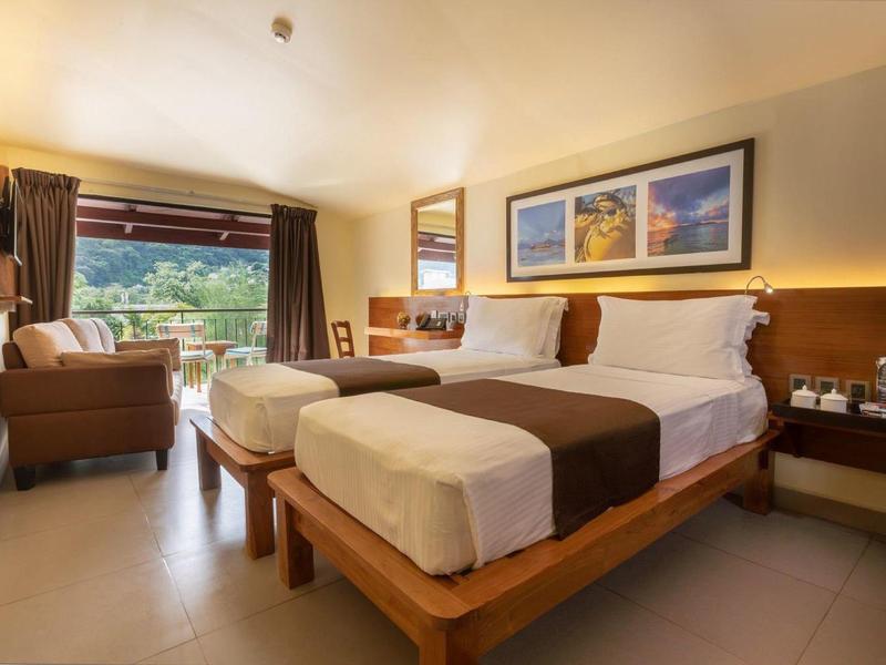 Habitación de hotel luminosa con dos camas, sillón y vista a un paisaje verde.