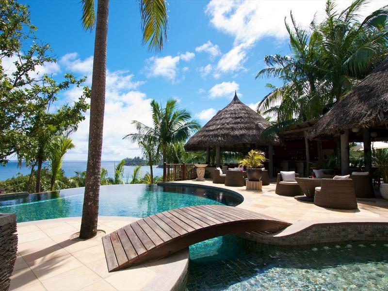 Area piscina di lusso con palme, lettini e vista mare sotto un cielo azzurro.