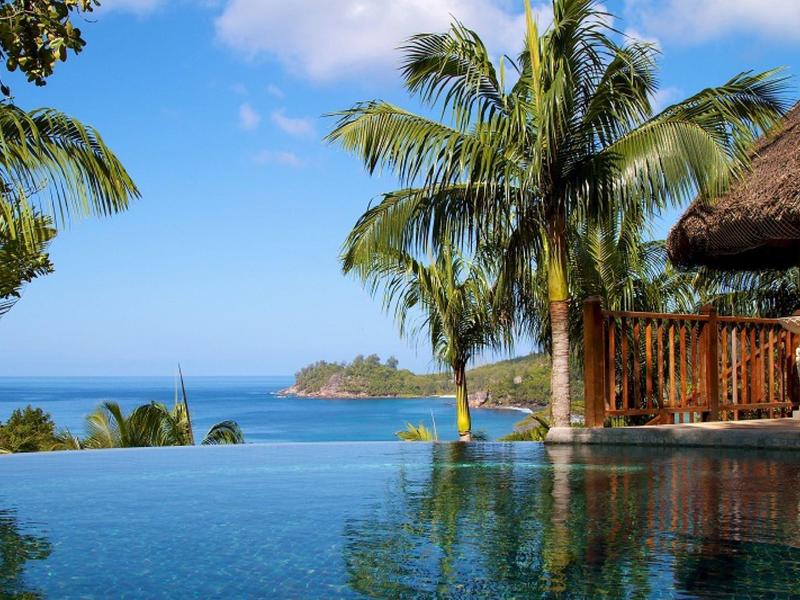Piscina a sfioro accanto a una capanna tropicale con palme e vista sul mare sullo sfondo.