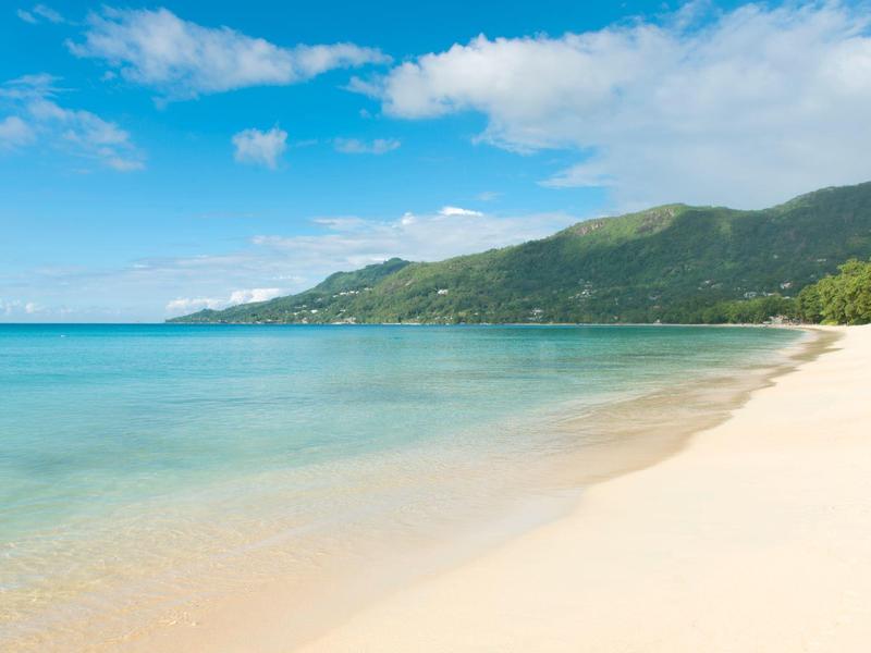 Weißer Sandstrand trifft auf ruhiges türkisblaues Meer mit grünen Hügeln unter blauem Himmel.