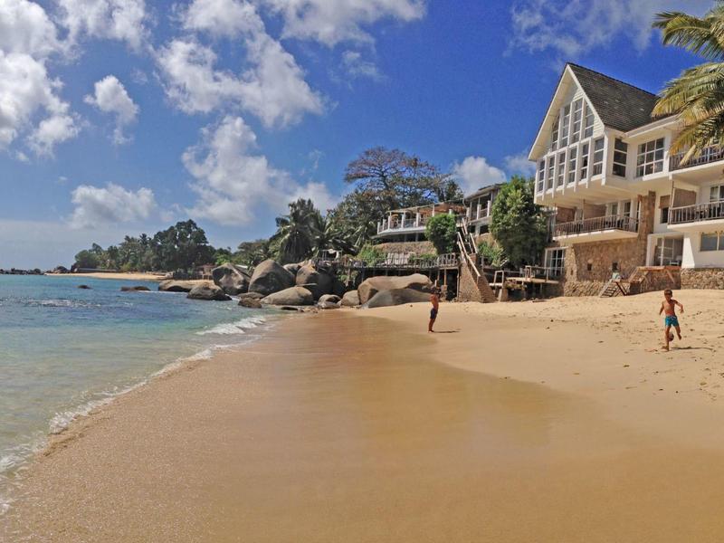 Sandstrand mit Palmen, Felsen und Häusern am Ufer, blauer Himmel mit weißen Wolken.
