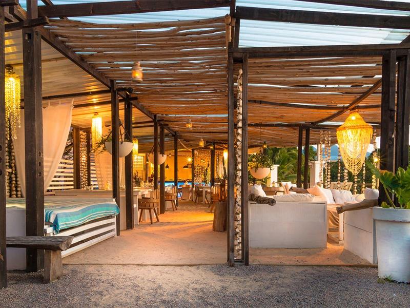 Gemütliche Außenlounge mit Holzdach, warmen Lichtern, Sitzmöbeln und Pflanzen bei Abenddämmerung.