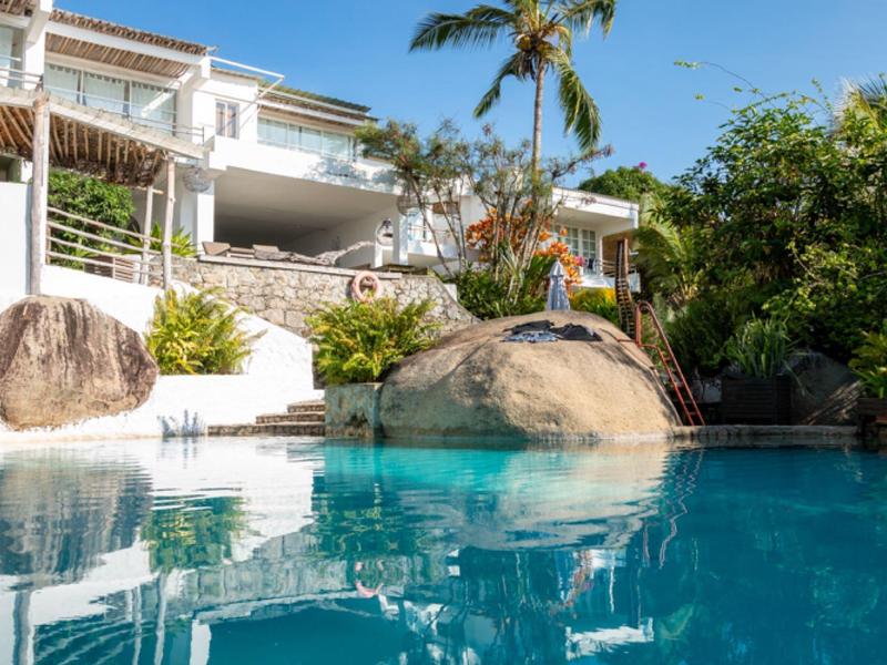 Moderner weißer Wohnhaus mit großem Pool, umgeben von Palmen, Felsen und grünem Garten bei sonnigem Himmel.