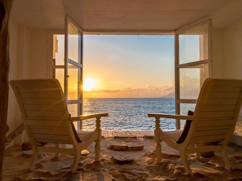 Zwei Holzstühle stehen auf Steinboden vor offenem Fenster mit Blick auf Meer und Sonnenuntergang.