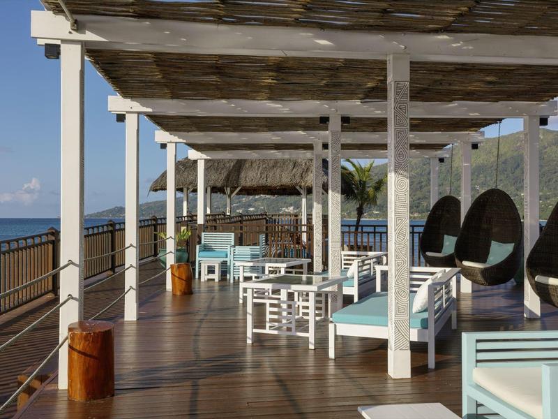 Offene Terrasse mit Holzfußboden, weißen Möbeln und Blick auf das Meer bei klarem Himmel.