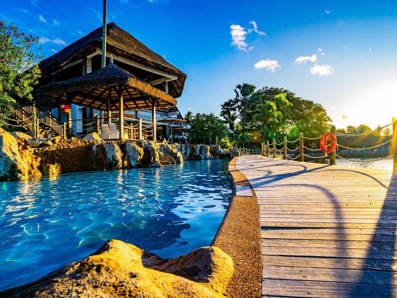 Ein sonniger Tag an einem Pool mit Holzsteg und einer Hütte umgeben von Bäumen.