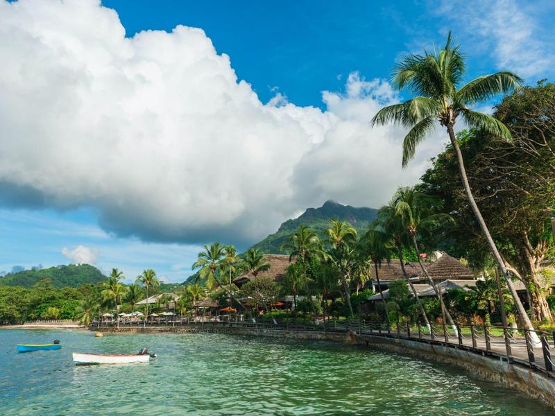 Klares Meer mit Ankern, Palmen und kleinen Hütten an der Küste unter blauem Himmel mit Wolken.